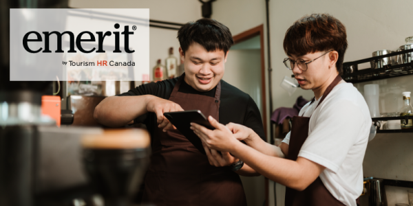 Two café employees wearing brown aprons are smiling and discussing an order while using a tablet. One employee is pointing at the screen while the other listens attentively. The 'emerit by Tourism HR Canada' logo is displayed in the top left corner.